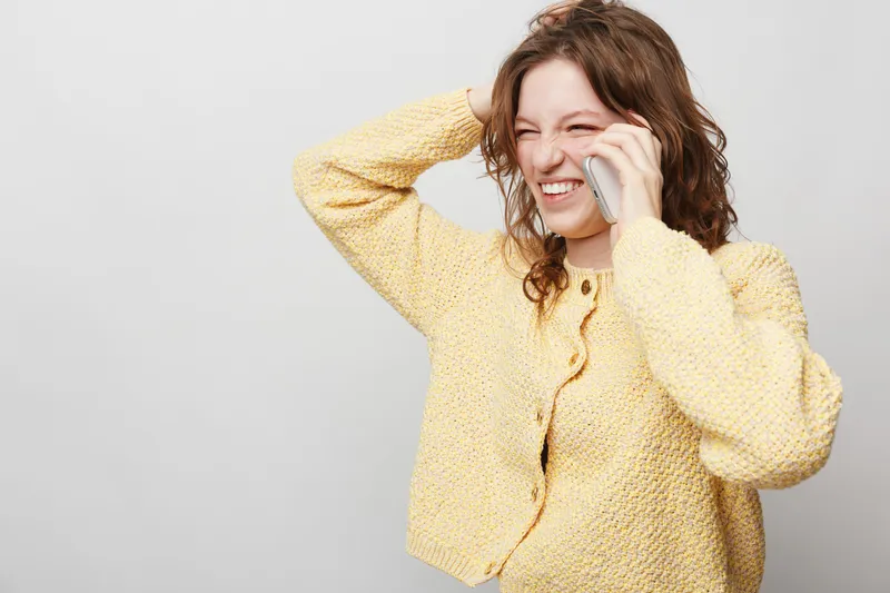Young woman in yellow sweater laughs while talking on phone, holding hand on head. Casual lifestyle, communication, happiness, and positive emotions concept on neutral background.