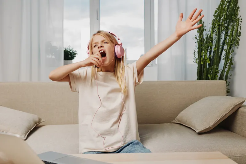 Happy girl wearing pink headphones singing and dancing on sofa in bright living room, enjoying music and carefree moment at home during daytime.