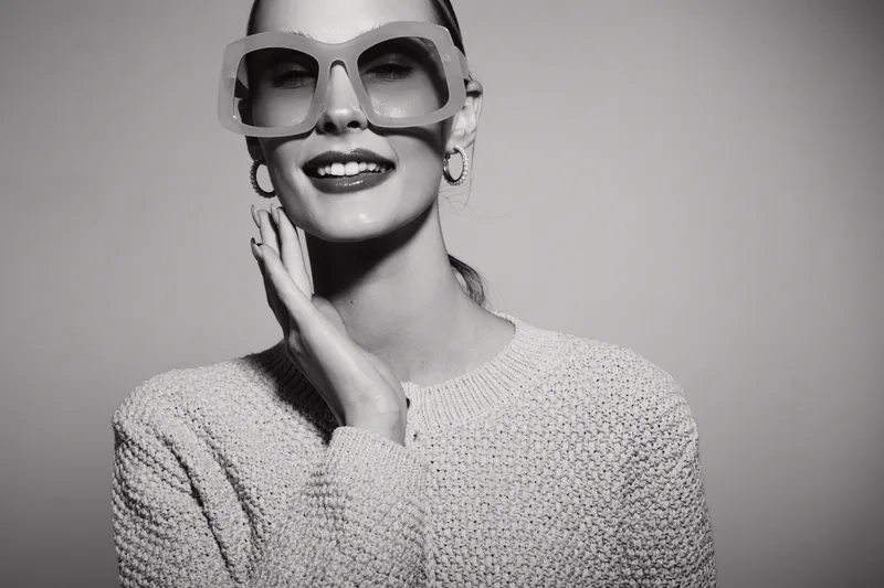 Stylish Caucasian woman with pale skin wearing oversized sunglasses and knitted sweater smiles confidently in studio lighting. Concept of fashion, beauty, and elegance.