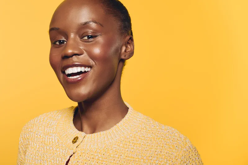 Happy smiling African woman with dark skin wearing yellow sweater stands against yellow background. Close-up portrait of joyful black female model with natural skin and bright mood.
