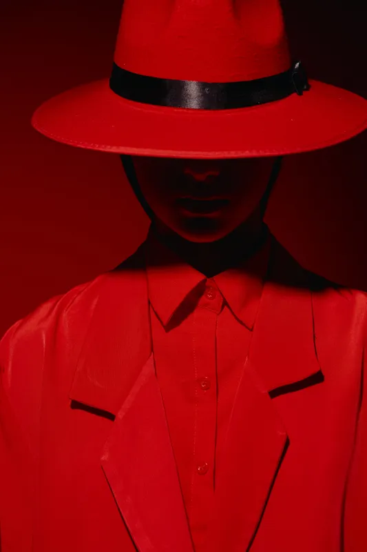 Person wearing vibrant red classic hat and matching sharp suit with black accents stands against red background. Fashion style portrait with strong creative lighting and shadows.