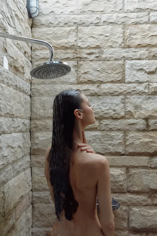 naked woman showering under rain showerhead in natural stone outdoor bathroom, wet hair cascading down her back, relaxed skin care moment