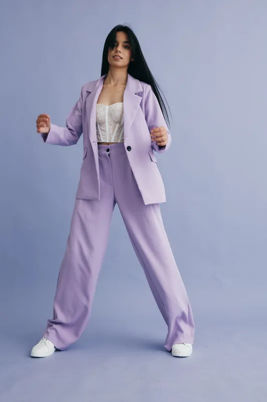 Stylish woman in lilac suit standing confidently against a soft purple background wearing white sneakers and lace top, showcasing modern fashion and elegance.