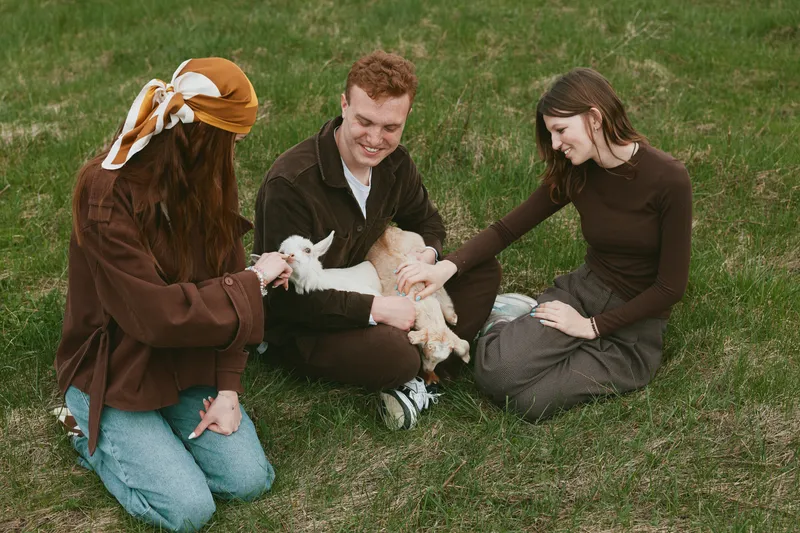Cheerful outdoors lifestyle scene with a group sitting on grass, a goat kid in the center, warm light, gentle interaction, relaxed smiles, casual outfits, wholesome bonding moment