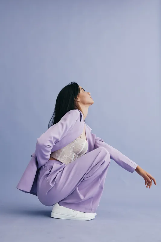 Woman posing in lilac suit with white sneakers, crouched down against lavender background, featuring modern fashion style and confident expression.