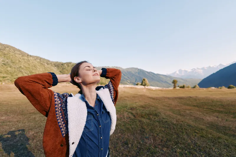 Woman nature mountains meadow relaxation outdoors travel freedom portrait of an adult in jacket and denim, eyes closed and arms behind head breathing fresh air in a serene valley landscape.
