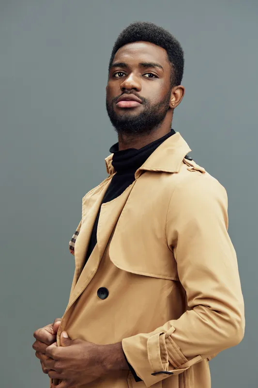Stylish African man in a beige trench coat poses confidently against a gray background, showcasing modern fashion and self assurance for contemporary lifestyle images