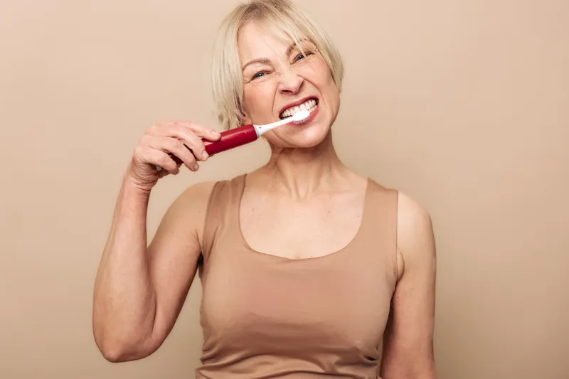 Blonde woman in beige top brushes teeth with electric toothbrush against neutral background. Healthy dental care, oral hygiene routine, and personal grooming concept with natural lighting.