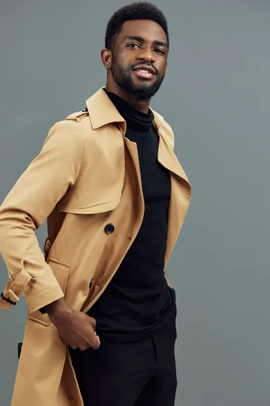 Smiling African man in stylish beige trench coat and black turtleneck, set against a muted gray background, perfect for fashion and lifestyle themes