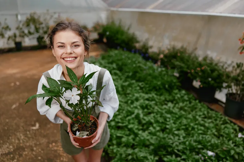 Happy young woman holding a potted plant in a greenhouse, showcasing her love for nature and gardening in a vibrant, lush setting