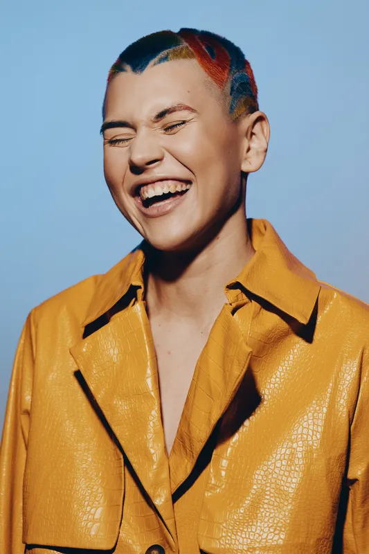 smiling woman with colorful short hair wearing a bright yellow jacket, joyful expression, studio background, vibrant and stylish portrait with playful energy