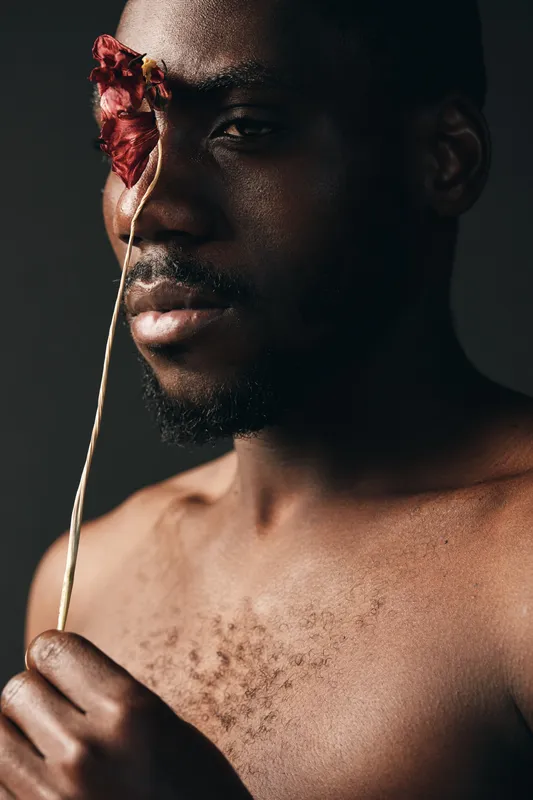 Creative conceptual portrait of African man with dark skin holding dried flower over eye in studio. Artistic visual style with closeup, bare chest and moody lighting for emotional expression.