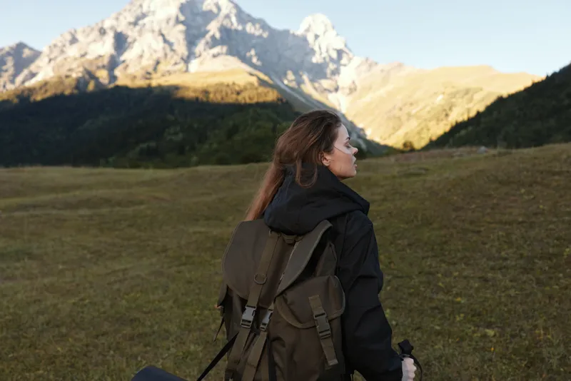 A lone traveler with a backpack stands in a grassy valley, looking toward towering mountains as soft light bathes the landscape, conveying adventure, exploration, and peaceful solitude.