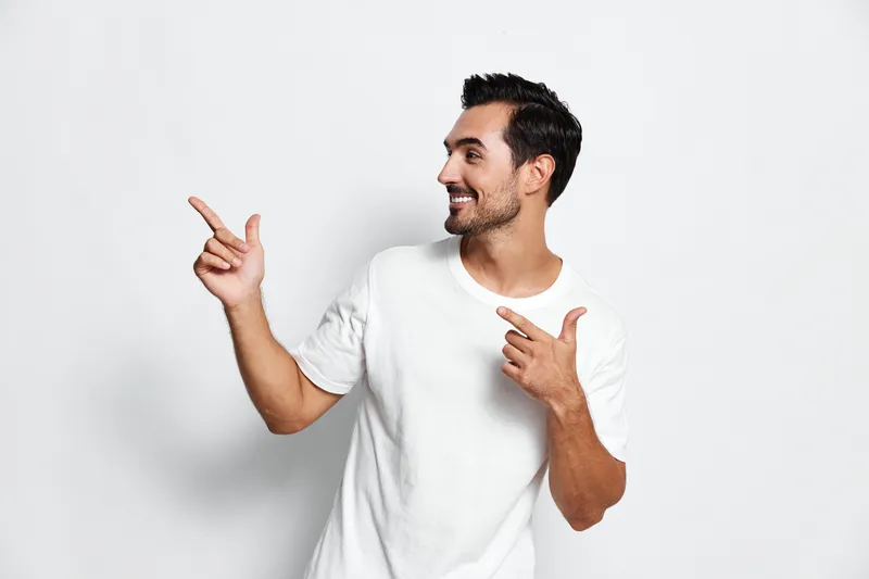 Smiling man in casual white t-shirt pointing with both hands, looking at the camera, exuding a cheerful and friendly vibe, isolated on plain solid white background.