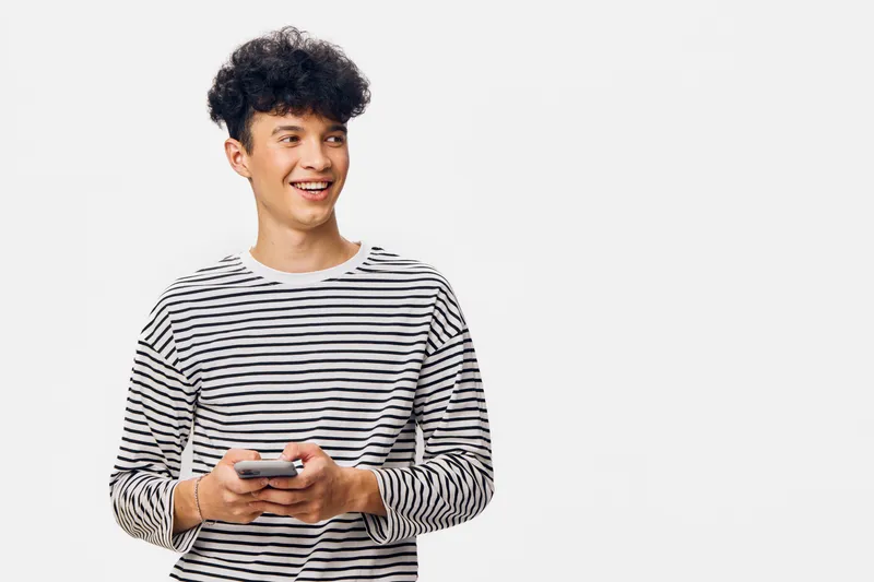 smiling young man in striped shirt holding smartphone, looking aside with cheerful expression against white background, casual lifestyle concept