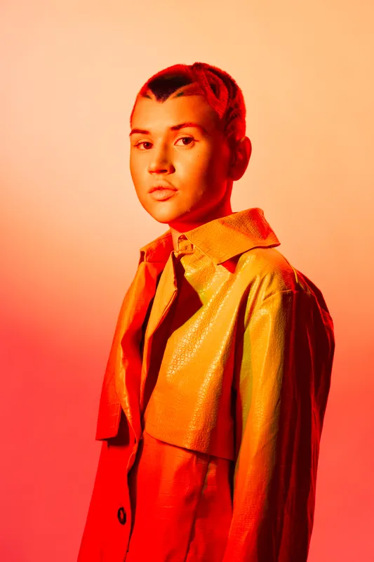 woman with short hair wearing shiny yellow jacket posing with serious expression against warm red and orange gradient background in studio lighting