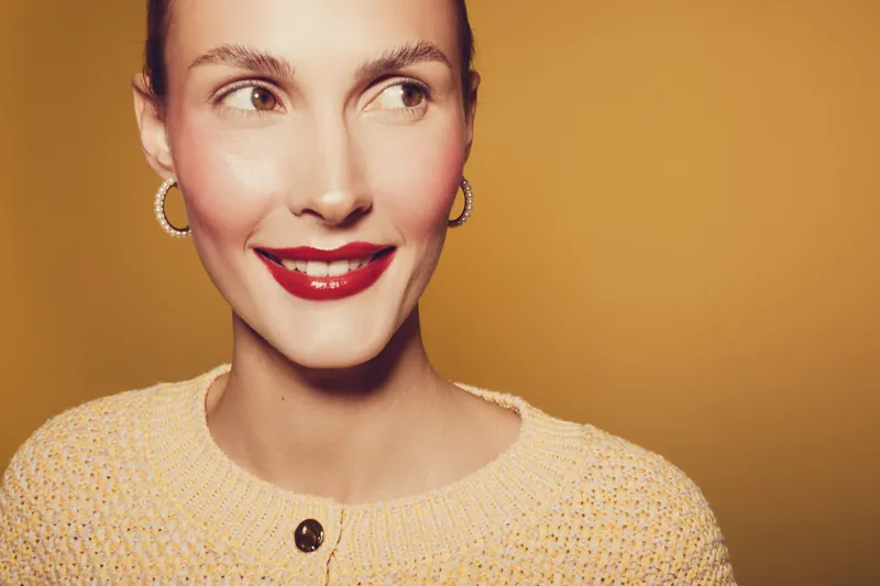 Smiling woman with red lipstick and hoop earrings looks sideways on yellow background. Close-up portrait with warm lighting and cozy sweater. Concept of beauty, fashion, and happiness.