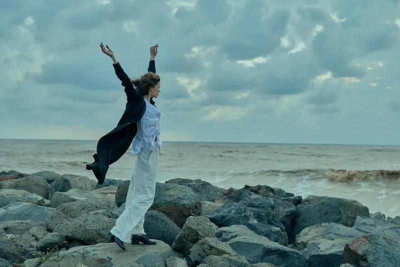 Woman on a rocky shoreline raises arms in motion, celebrating outdoors with sea spray and dramatic clouds, capturing energy, resilience and exploration in a dynamic, cinematic moment.