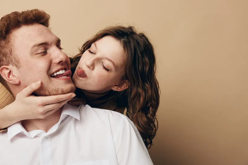 Playful young woman with curly hair teasing smiling man with red hair and beard. Couple in casual clothes share joyful moment on beige background. Concept of love, fun, and relationship.
