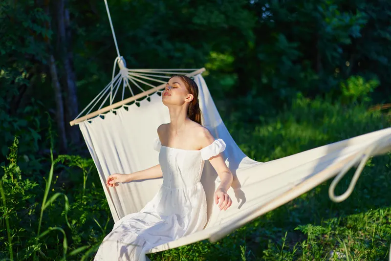 Young woman enjoying relaxation in a hammock surrounded by lush greenery, wearing a white summer dress, exuding tranquility and joy in a serene outdoor setting