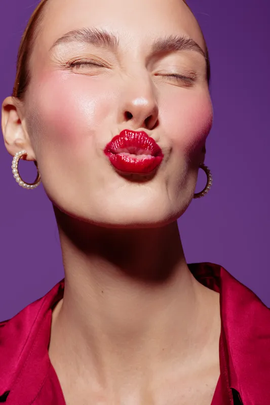 Close-up of a woman with red lipstick puckering lips and closed eyes against purple background. Concept of beauty, makeup, fashion, and self-expression in studio lighting.