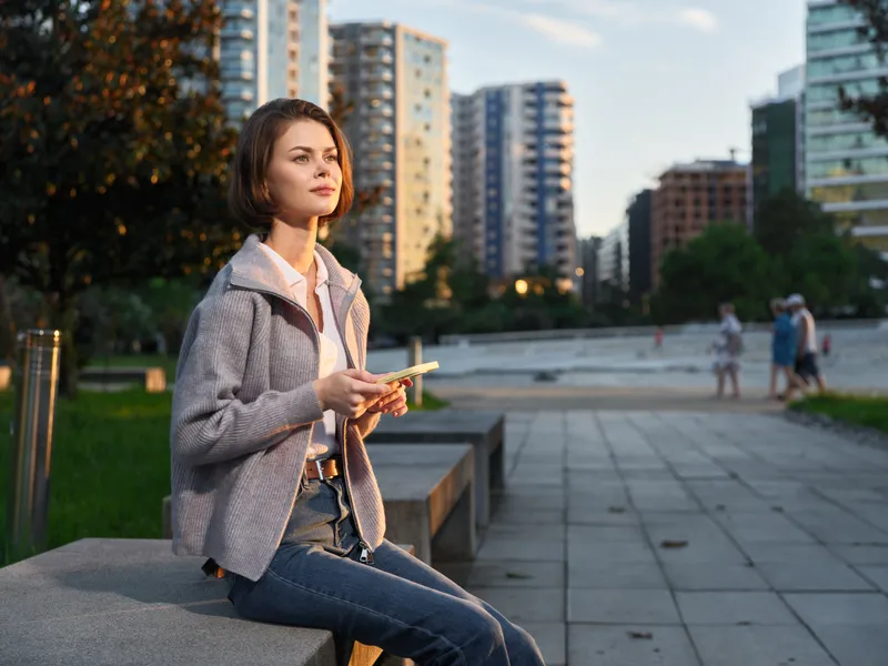 A focused young woman sits on a concrete bench in an urban park, using a tablet. She wears a light cardigan and jeans, with modern high rise buildings in the background, conveying a relaxed urban