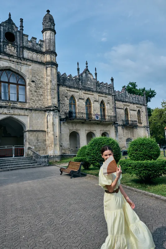 A woman in a flowing dress stands outdoors in front of a historic castle, holding a book and posing gracefully amid manicured hedges and stone architecture.