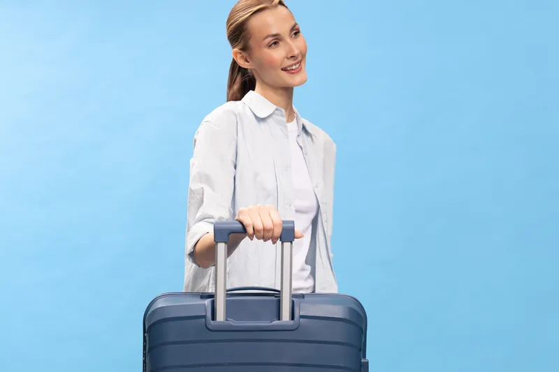 Smiling young woman in casual clothes holds blue suitcase handle, standing against light blue background. Concept of travel, vacation, and holiday preparation with positive mood.