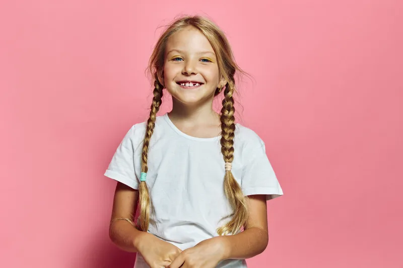 Smiling girl with two braids wears a white t-shirt standing against a pink background. She shows missing front teeth and looks directly at the camera.