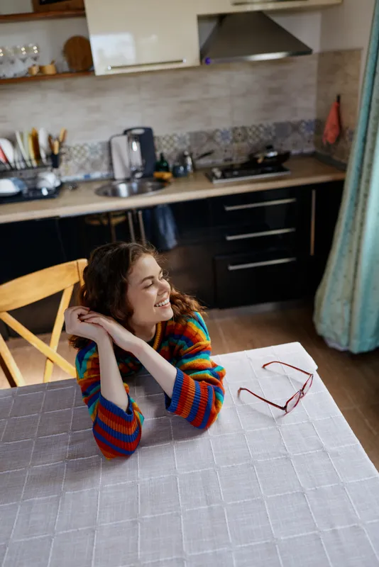 Cheerful woman sits at a kitchen table in a striped sweater, enjoying a relaxed moment at home, a candid lifestyle scene with warm light, cozy mood, and content expression