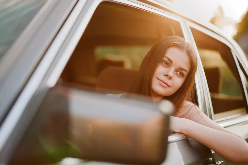 Young woman with long hair enjoying a sunset view from a car window, showcasing a thoughtful expression and casual summer attire.