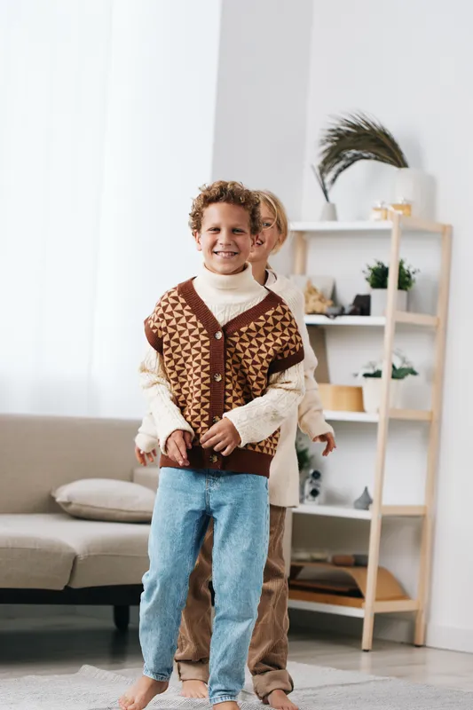 Two children stand indoors, one in a patterned knitted cardigan and cozy sweater, smiling warmly as they hug, creating a sweet moment of friendship and comfort in a living room.