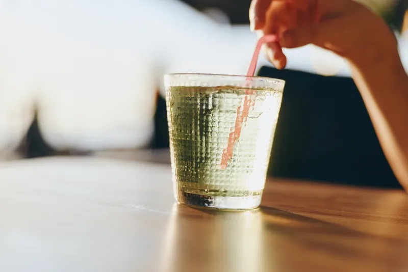 glass of sparkling water with hand stirring pink straw on wooden table in bright natural light, refreshing summer drink