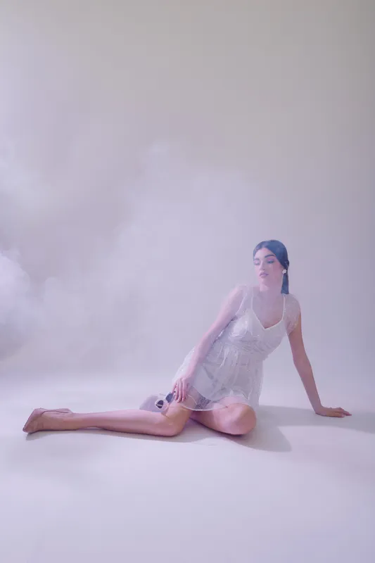 Young woman in a white dress poses on the floor in a soft pink studio with light smoke. Concept of beauty, fashion, elegance, and delicate feminine style.
