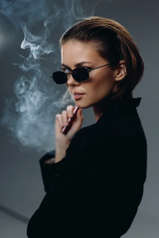 Stylish woman wearing sunglasses smoking with rising smoke against dark background