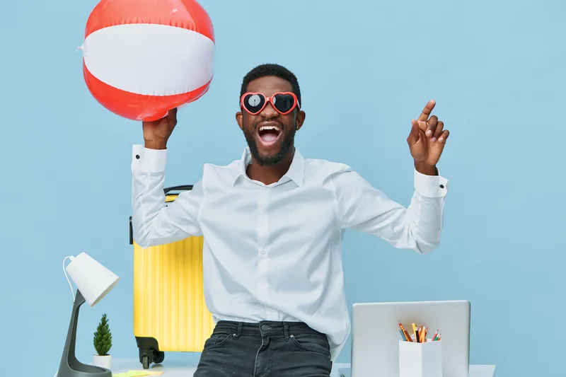 Joyful man celebrating summer vacation holding striped beach ball, wearing sunglasses and casual white shirt in modern colorful office with travel accessories. Happiness, leisure, relaxation concept.