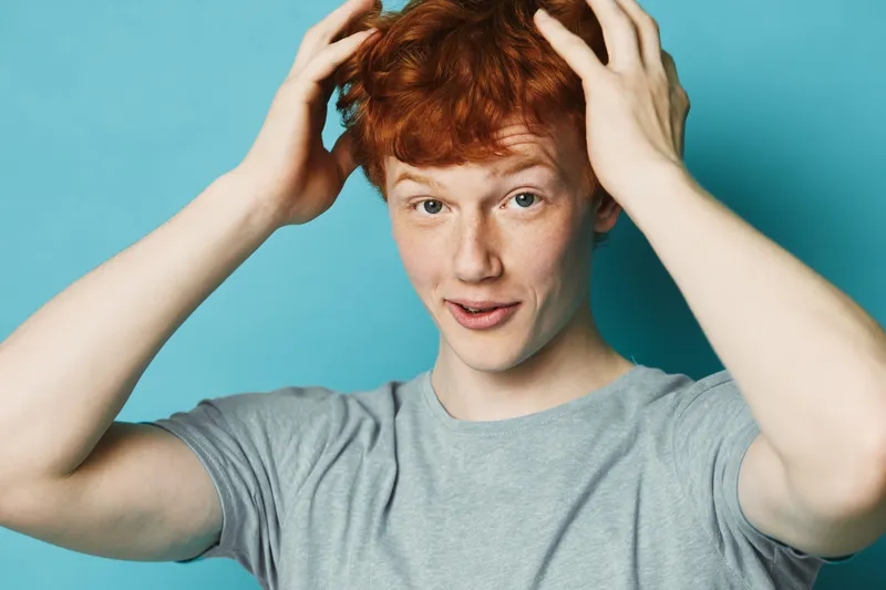 Young man with red hair and freckles touching his head with hands, expression of surprise or realization, wearing grey t shirt against blue background.