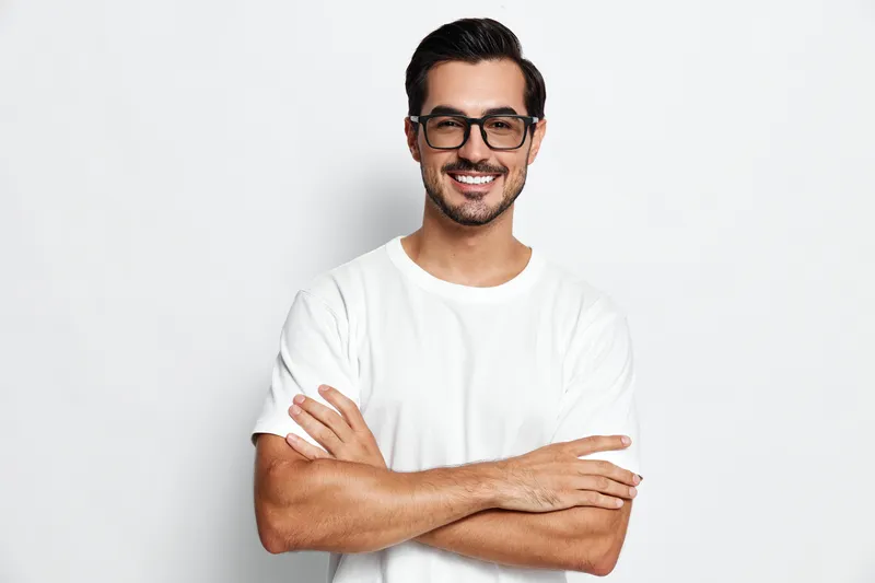 Smiling man wearing glasses and white t-shirt stands confidently with crossed arms against a solid background. Portrait reflecting positive vibes and casual style.
