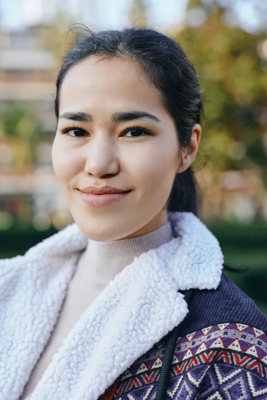 Woman with a warm smile in a cozy fleece and patterned outerwear, standing outdoors with a soft blurred background, showcasing casual style, natural charm and daytime light in a candid fashion moment