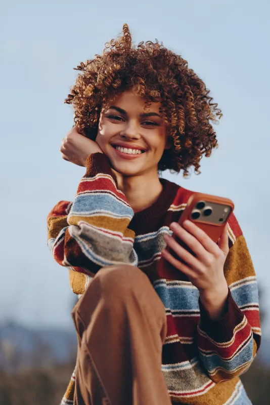 Woman lifestyle portrait of a smiling curly haired woman outdoors, holding a smartphone and adjusting hair, casual knit sweater in daylight, friendly mood for mobile communication