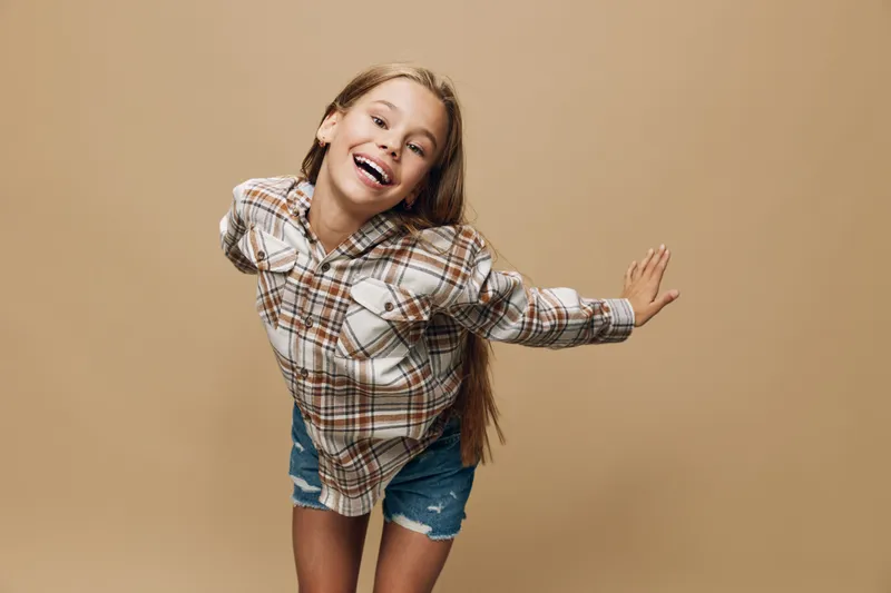 Smiling girl with long hair wearing casual plaid shirt and denim shorts posing playfully on beige background. Happy child in relaxed youth fashion with joyful expression.