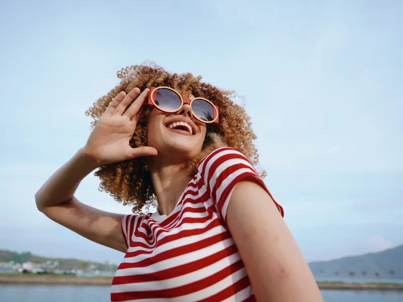 Joyful woman with curly hair wearing a red and white striped shirt and round sunglasses smiles outdoors against a bright blue sky, capturing carefree summer vibe.
