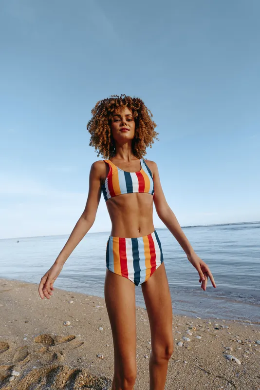 Woman beach ocean swimsuit striped curly hair walking summer sunlight outdoors, confident relaxed posture, blue sky horizon and gentle waves behind, soft sea breeze, natural colors, lifestyle fashion