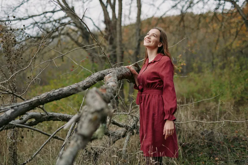 A woman in a red dress stands beside a fallen tree in an autumn meadow, smiling upward with a sense of genuine presence and authenticity, capturing a credible, natural lifestyle moment.