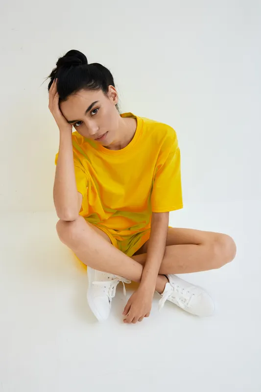 Young woman in bright yellow t-shirt sitting on the floor with hand on head, expressing thoughtful mood against a clean white background, casual style outfit.