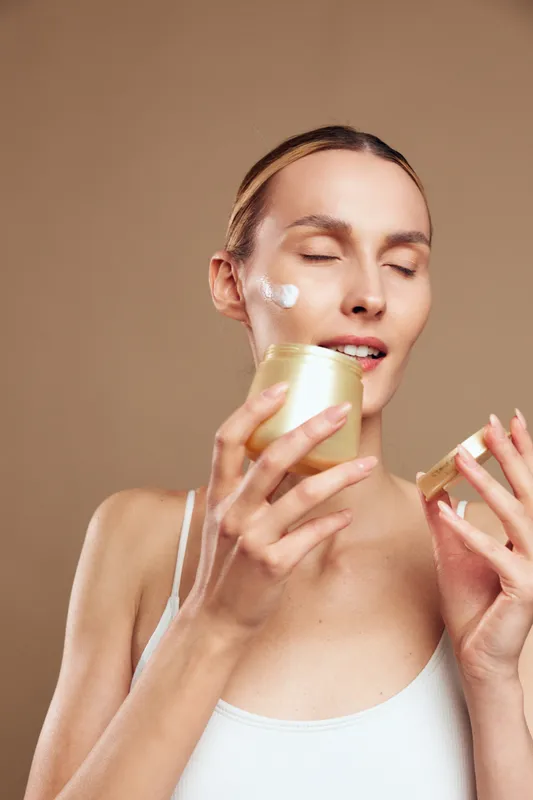 Young woman applying facial cream on cheek with closed eyes, skincare routine, natural beauty, soft lighting, neutral background, healthy glowing skin, self care moment