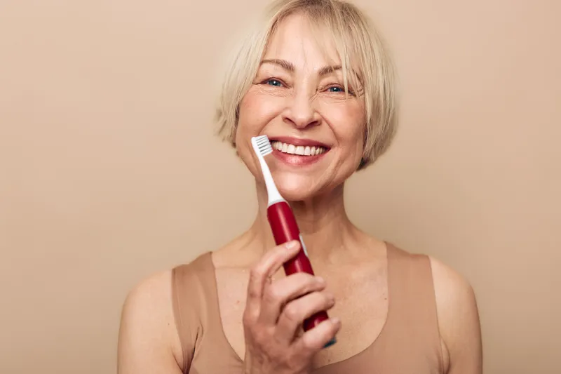 Smiling mature woman with short blonde hair holding electric toothbrush near face. Close-up on beige background. Concept of dental care, oral hygiene, and healthy lifestyle for seniors.