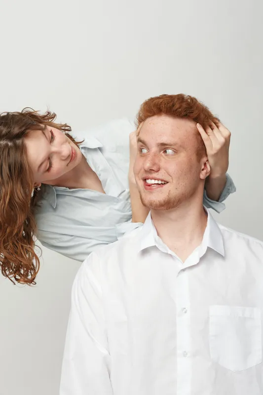 Young redhead man in white shirt smiles while woman with curly hair touches his head gently. Studio portrait on light background. Concept of affection, friendship, and casual interaction.