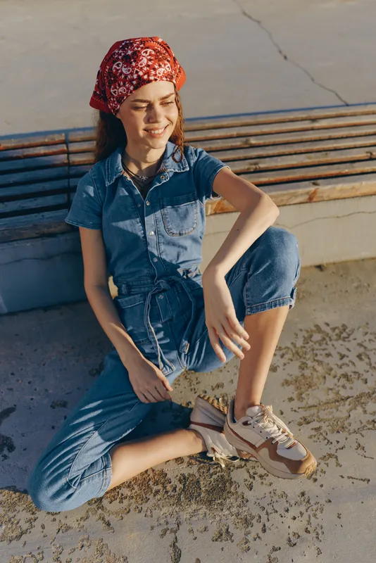 Lifestyle woman wearing modern boho-western denim street style, smiling with red bandana and casual sneakers in warm film color lighting outdoors.