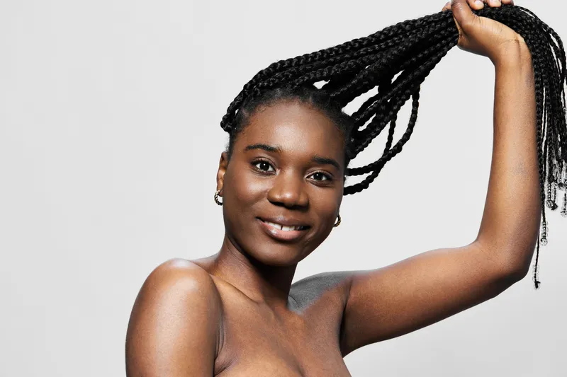 Young Black woman with braided hair playfully holding her hair, showcasing confidence and beauty against a minimalistic gray background